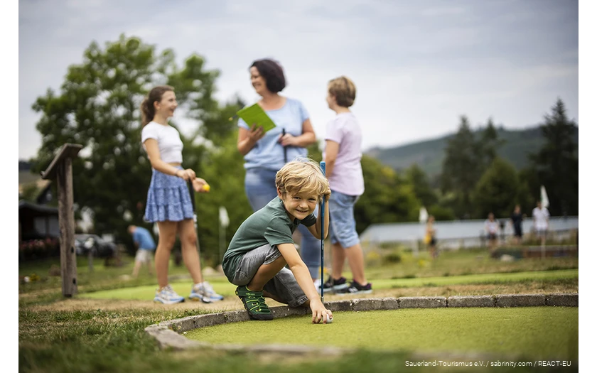 A family playing mini golf at Diemelsee. A son prepares his next shot.