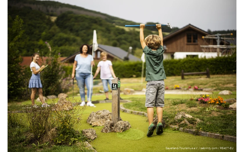 A family playing mini golf. In the foreground, a son is happy and jumps into the air.