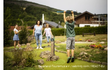 Eine Familie spielt Minigolf. Im Vordergrund freut sich ein Sohn und springt in die Luft.