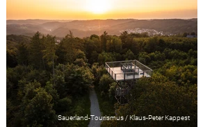Blick von oben auf den Turm. Im Hintergrund geht die Sonne unter und man sieht verschiedene Ortschaften