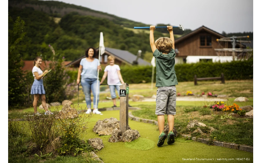 Een gezin speelt minigolf. Op de voorgrond is een zoon blij en springt in de lucht.