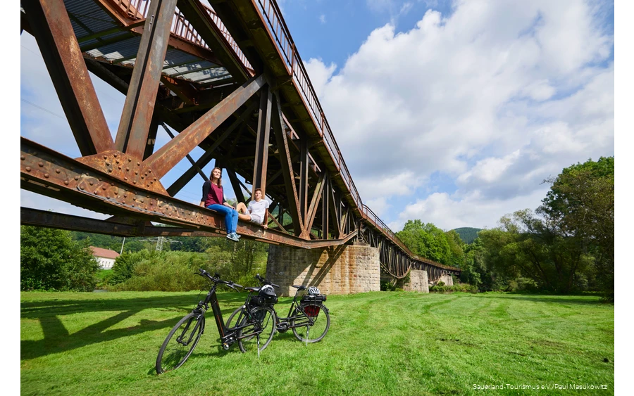 Pärchen sitzt auf dem Träger einer Brücke unter ihnen zwei E-Bikes