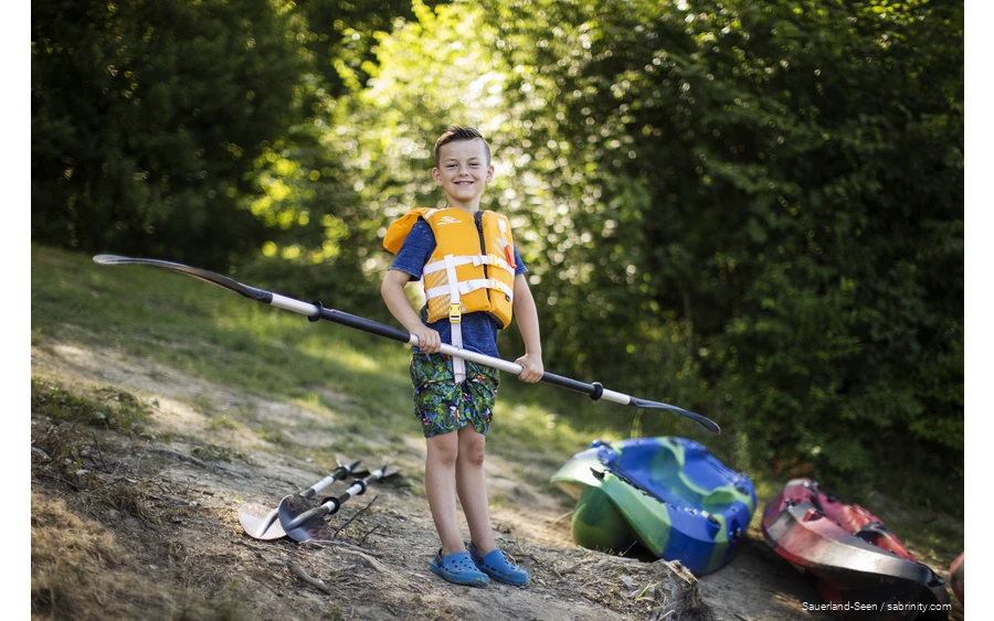 Junge mit Schwimmweste freut sich auf Kanufahrt mit einem Paddel in der Hand.