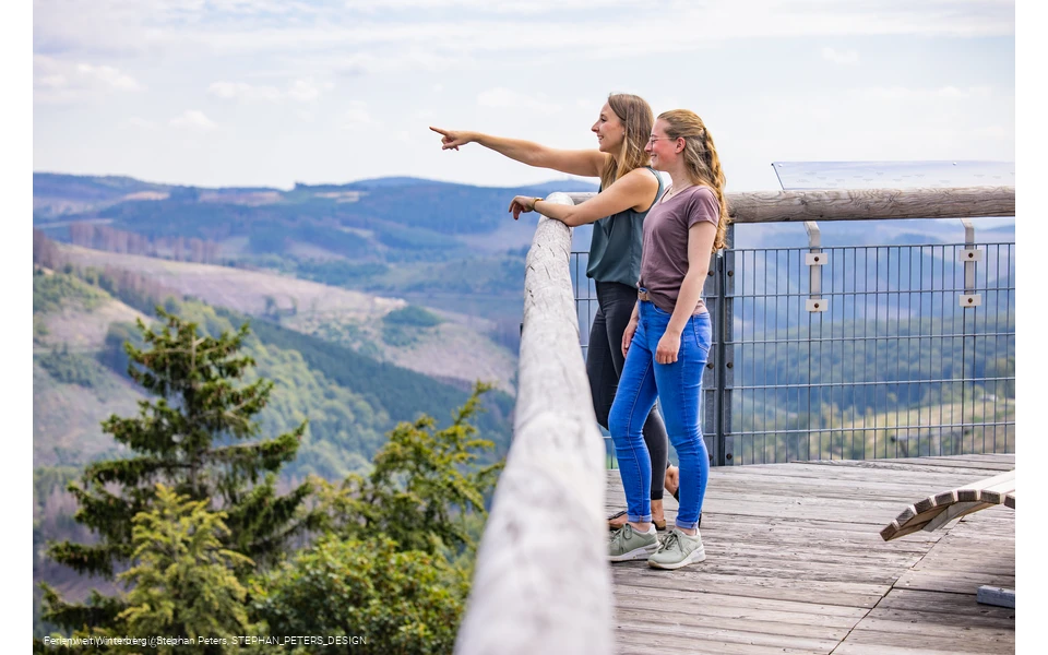 Zwei Frauen stehen auf einer hohen Brücke und blicken in die Ferne.