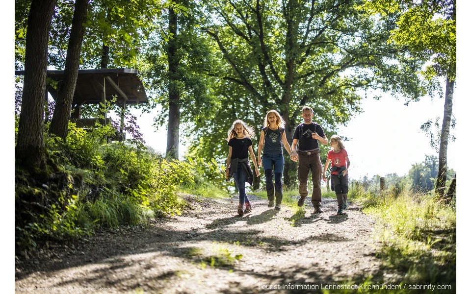 Eine vierköpfige Familie wandert fröhlich und beschwingt einen Wanderweg entlang. Die Eltern halten Händchen, alle lachen.