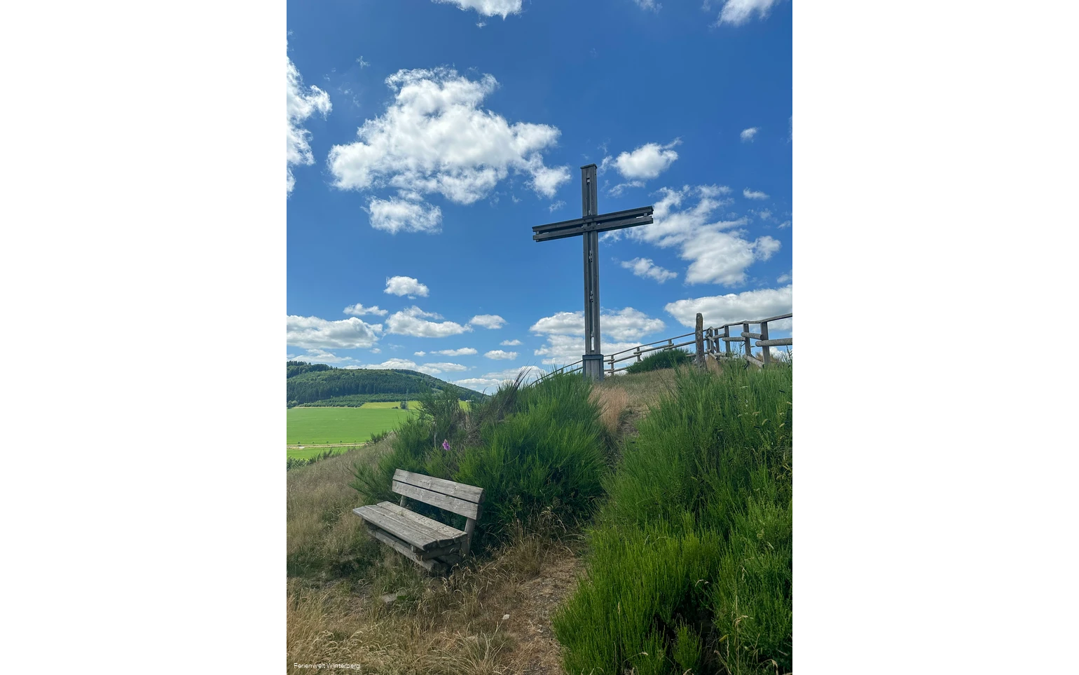 Ein großes Holzkreuz auf einem begrünten Hügel, davor eine Bank.