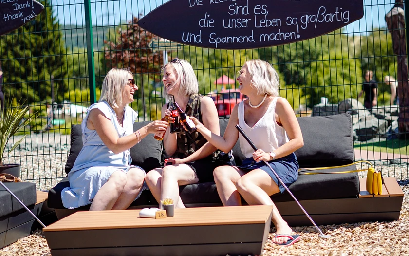Drei Frauen sitzen auf einer Bank am Tisch und stoßen mit Gläsern an.