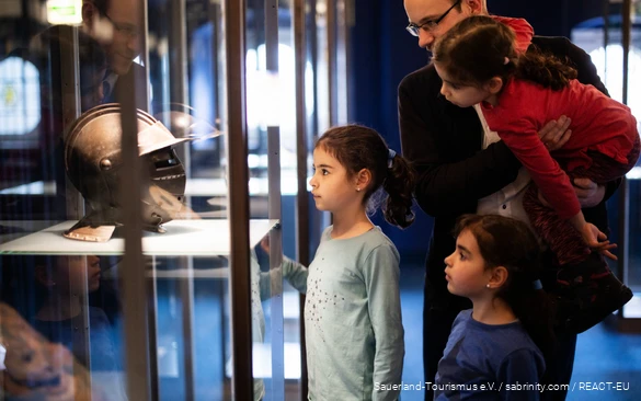 Vater steht mit drei Töchtern vor einer Vitrine in einem Museum. Sie schauen sich einen Ritterhelm an.
