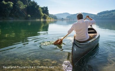 Ein Mann in einem weißen Hemd  sitzt in einem Kanu auf dem Hennesee und die Sonne spiegelt sich im Wasser