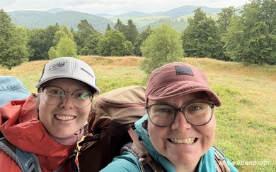 Anne Abendroth and her friend grin into the camera on one of their hikes. They are wearing outdoor clothing, Kappe hats and hiking rucksacks.