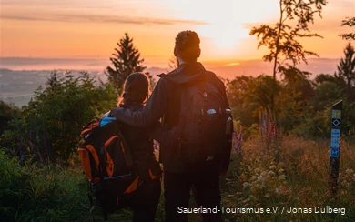 Man en vrouw staan arm in arm met hun rug naar de camera en kijken naar een kleurrijke zonsondergang vanaf een uitkijkpunt. Ze dragen rugzakken en dragen outdoorkleding.