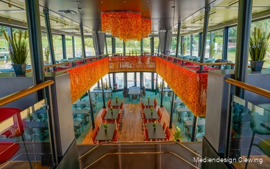 Innenansicht der EMS Hennesee mit Blick auf zwei Decks, zahlreichen Tischen und warmem orangenen Dekor.