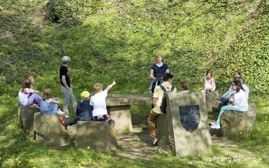 Kinder sitzen auf Steinen in einem Halbkreis in Richtung der Gruppenführerin. Ein Junge zeigt auf.