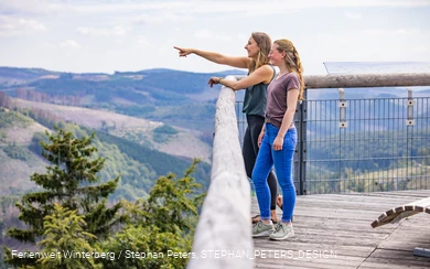 Zwei Frauen stehen auf einer hohen Brücke und blicken in die Ferne.