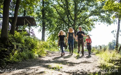 Eine vierköpfige Familie wandert fröhlich und beschwingt einen Wanderweg entlang. Die Eltern halten Händchen, alle lachen.