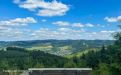 Bank auf einem Felsen mit weiter Aussicht über Wiesen und Wälder und einer Ansammlung von Häusern.