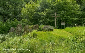 Eine Ansammlung von großen Steinen neben einer Bank und einem großen Holzkreuz, sowie einer alten hölzernen Tafel mit Informationen zu den Opfersteinen im Hohlen Graben.