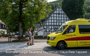 Ein Wanderpärchen winkt einem gelben Mini-Bus, der vor einem Fachwerkhaus geparkt hat.