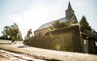 Zwei Rennradfahrer fahren um eine Kurve an einer Kirche vorbei.