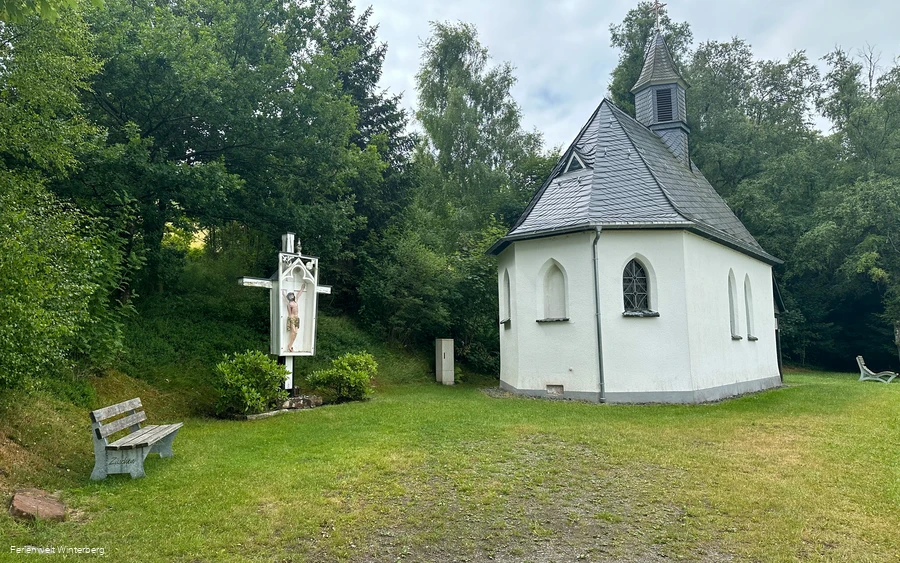 Kapelle mit einer Jesusfigur am Kreuz und einer Bank, umgeben von Bäumen.