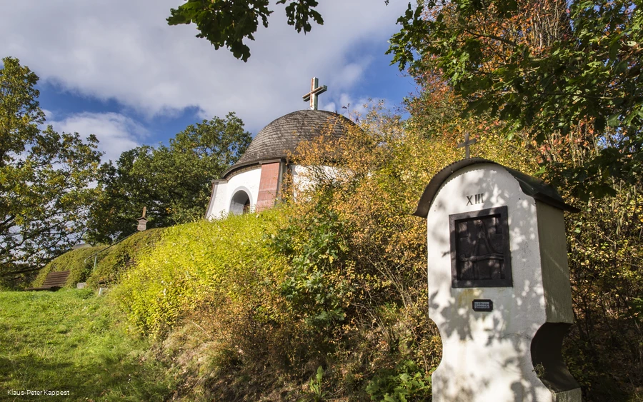Eine Station eines Kreuzweges mit einem Teil einer Kapelle im Hintergrund umgeben von Wiesen und Bäumen.
