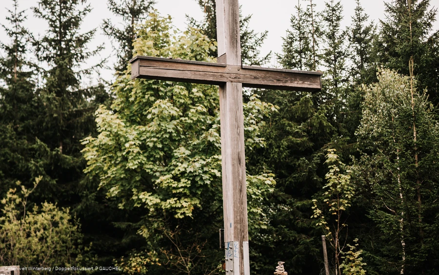 Ein großes Holzkreuz auf einer Wiese mit Wald im Hintergrund.