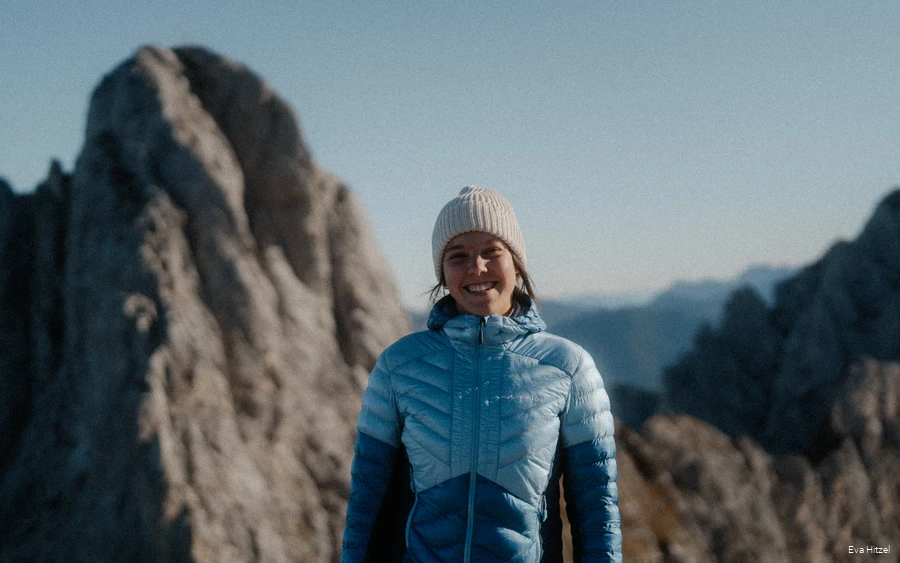 Eva von Wildwärts Eva vom Blog Wildwärts grinst mit blauer Daunenjacke und Mütze bekleidet in die Kamera. Im Hinzergrund sind schroffe Berge zu sehen.