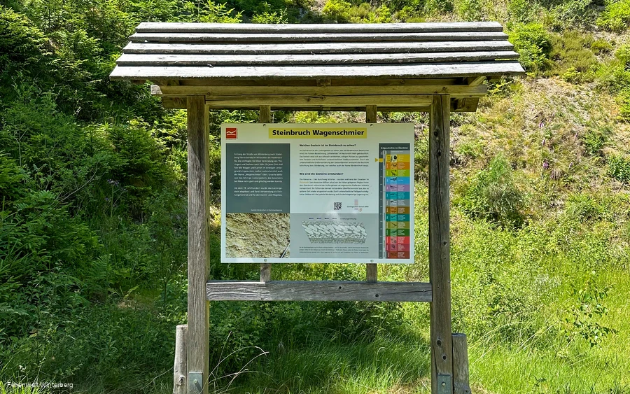 Informationstafel zum Steinbruch Wagenschmier mit grün bewachsenem Steinbruch im Hintergrund.
