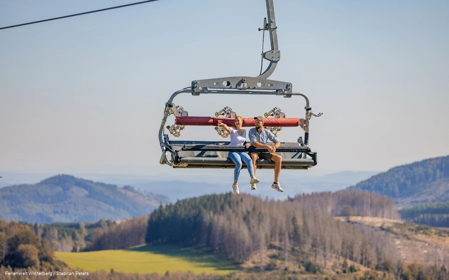 Zwei Personen sitzen in einem Sessellift mit einer Berglandschaft im Hintergrund.