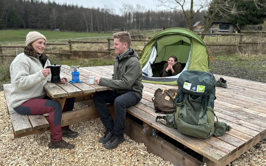 Two hikers are sitting at a table at a trekking site in the Arsnberg Forest. In the background is a tent with a woman looking out of it.