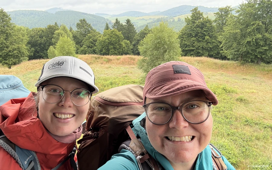 Anne Abendroth and her friend grin into the camera on one of their hikes. They are wearing outdoor clothing, Kappe hats and hiking rucksacks.