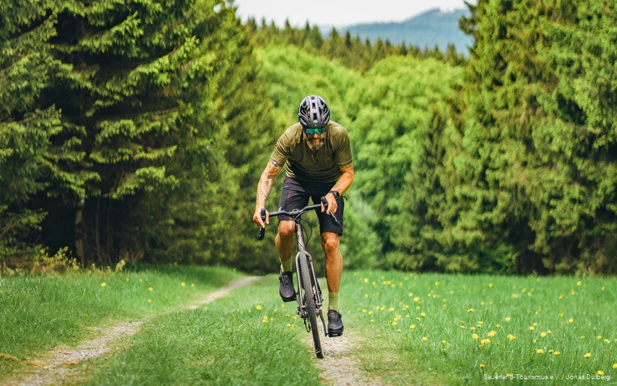 Gravelstory 2 Tätowierter Mann auf einem Gravelbike von vorne. Er fährt einen kleinen Pfad umgeben von Nadelbäumen hoch.