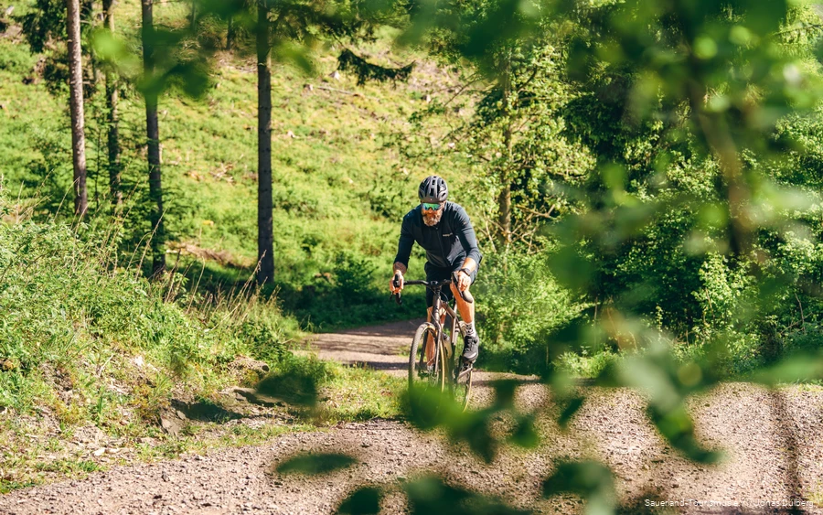 Gravelstory 3 Tätowierter Mann auf einem Gravelbike von vorne. Er fährt einen Schotterweg hoch.