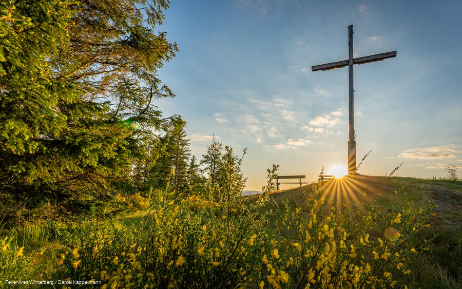 Ein großes Holzkreuz auf einem Hügel neben einer Bank vor blauem Himmel und Sonne.