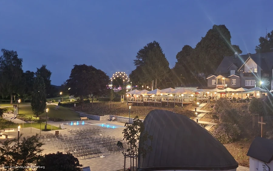 Drohnenaufnahme des beleuchteten Restaurants Kurparkstuben Korn mit dem beleuchteten Hotel Oversum im Hintergrund und der Musikmuschel mit Sitzmöglichkeiten im Vordergrund.