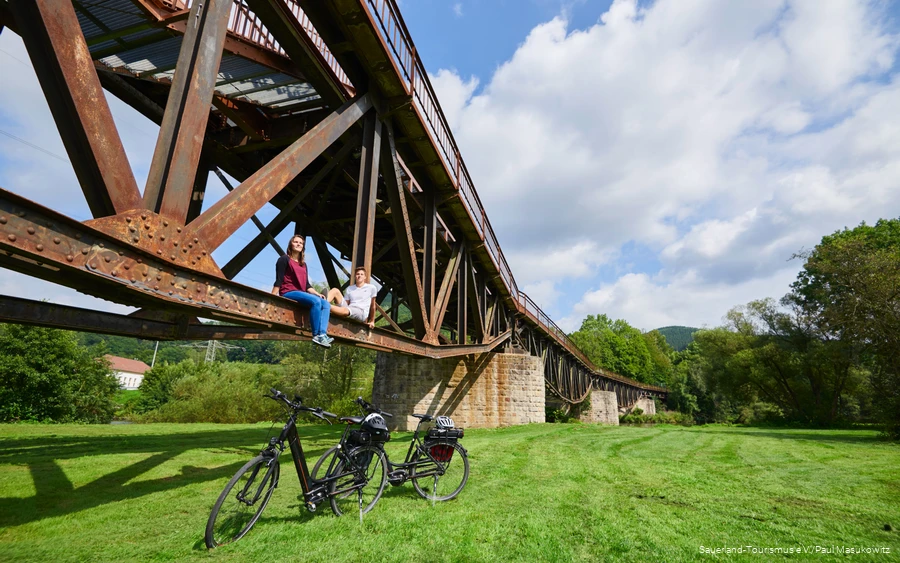 Pärchen sitzt auf dem Träger einer Brücke unter ihnen zwei E-Bikes