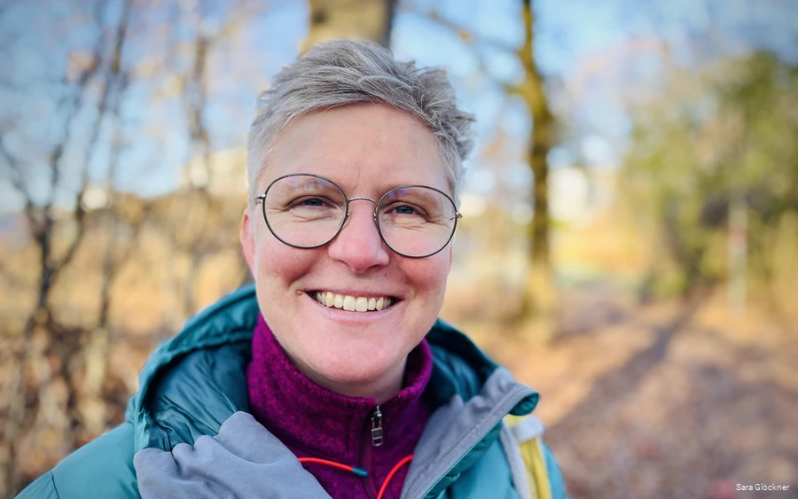 Sara Glöckner trägt Outdoorkleidung und grinst in die Kamera. Sie steht im Wald, den man verschwommen im Hintergrund sehen kann.