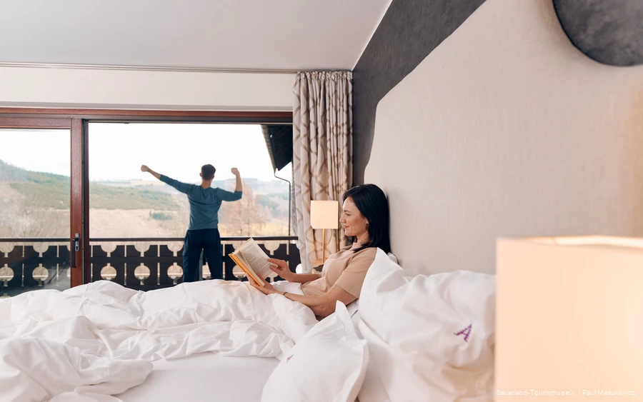 Eine Frau liegt im Hotelbett und liest entspannt ein Buch, während ihr Mann hinter ihr auf dem Balkon steht und sich streckt.