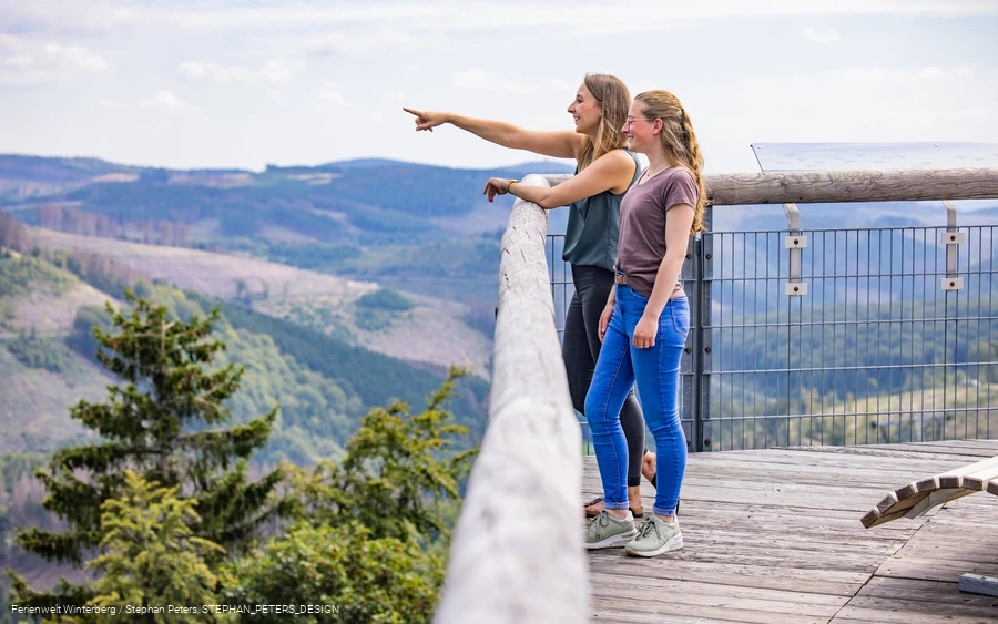 Zwei Frauen stehen auf einer hohen Brücke und blicken in die Ferne.