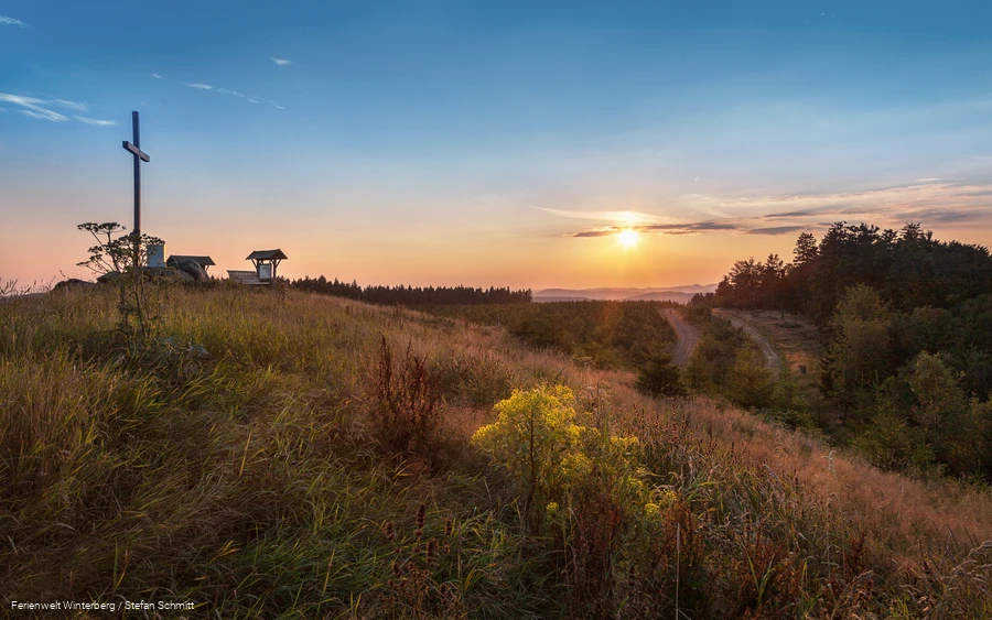 Ein großes Holzkreuz auf einem Hügel, umgeben von Heidelandschaft mit der aufgehenden Sonne im Hintergrund.