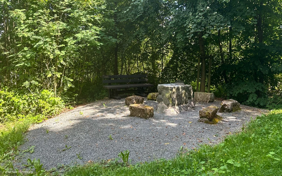 Einige große Steine, welche im Kreis um einen großen Steinblock angelegt sind, umgeben von Wiese und Bäumen.