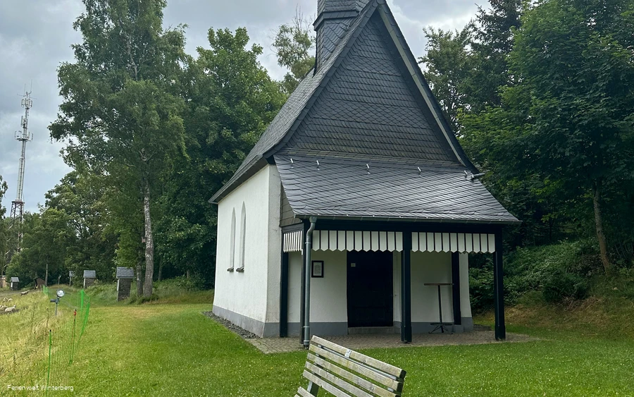 Bank vor einer Kapelle, welche von Bäumen umgeben wird.