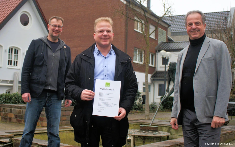 Three men smile, the middle one holds the Sauerland Tourism membership certificate