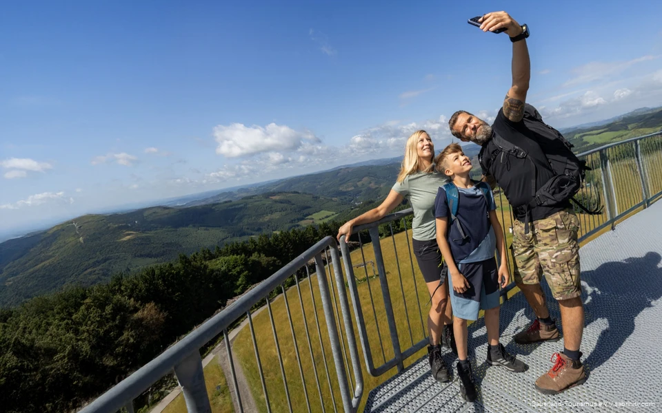 Eltern mit Sohn stehen auf einem Aussichtspunkt und machen ein Selfie mit dem Ausblick auf die sauerländer Berge im Hintergrund.