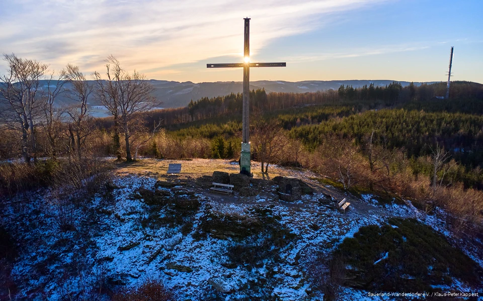 Landscape shot of the summit cross on the Olsberg at sunrise. There is a thin layer of snow.