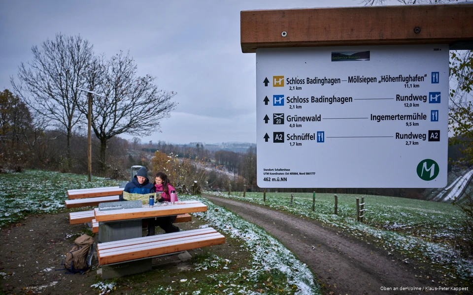 Op de voorgrond staat een wegwijzer, op de achtergrond zit een stel op een bankje naar een kaart te kijken. Er ligt een licht laagje sneeuw.