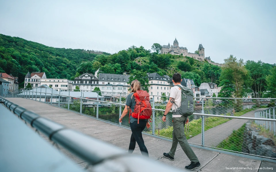 Zwei junge Wanderer überqueren eine Brücke und blicken zur Burg Altena hinauf.