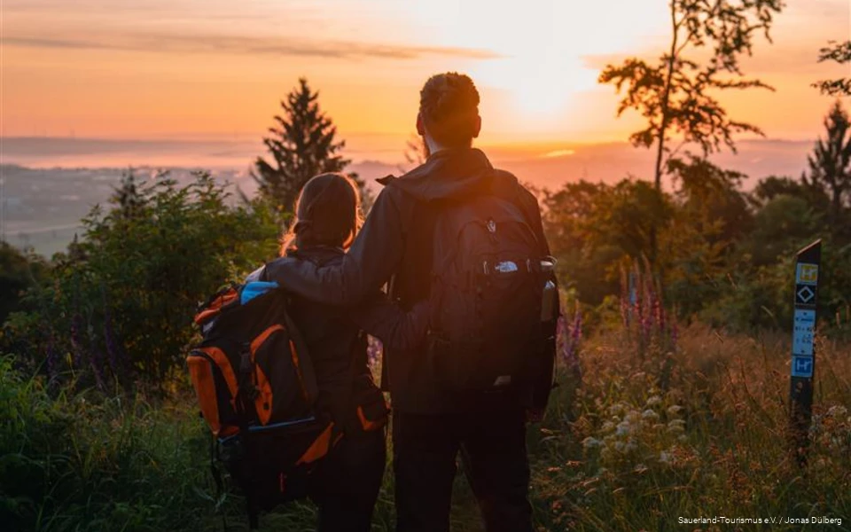 Man en vrouw staan arm in arm met hun rug naar de camera en kijken naar een kleurrijke zonsondergang vanaf een uitkijkpunt. Ze dragen rugzakken en dragen outdoorkleding.