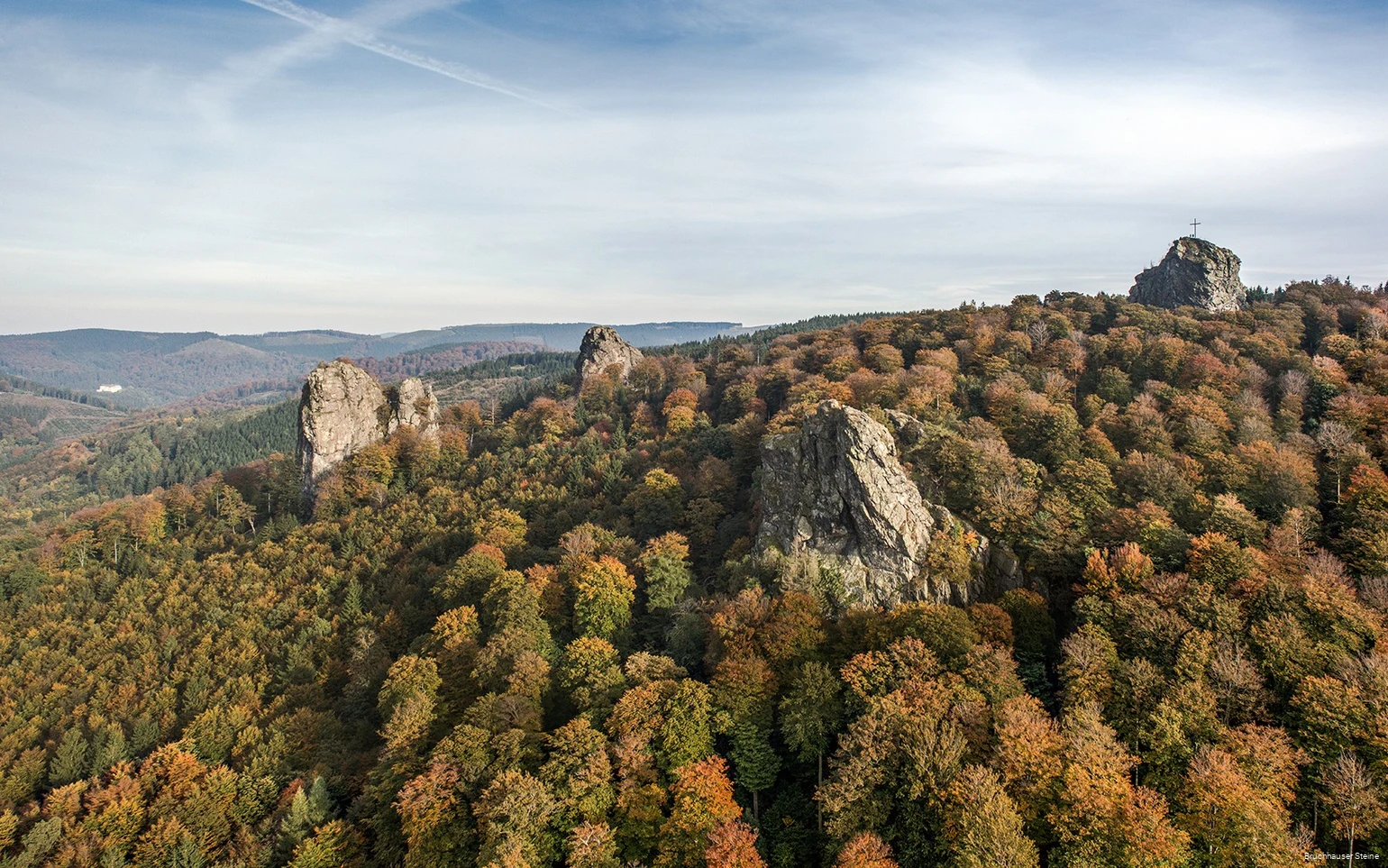 Luftaufnahme der Bruchhauser Steine, die zwischen herbstlich verfärbtem Laub hervorragen. Auf dem rechten Stein steht ein Gipfelkreuz.