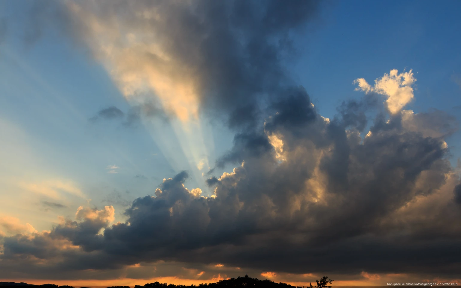Dunkle Wolken am Abendhimmel mit orangenem Horizont und Sonnenstrahlen, die hinter den Wolken hervorkommen.